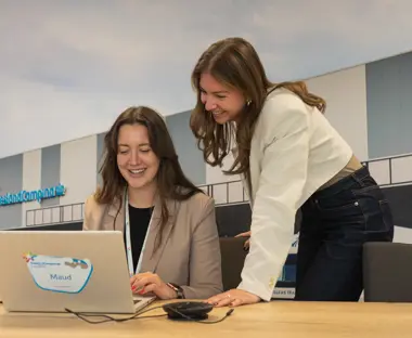 Twee jonge vrouwen in een kantoorruimte, lachend achter een laptop met het FrieslandCampina-logo en de naam Maud op een sticker; op de achtergrond een fotowand van een distributiecentrum.
